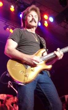 Johnston playing a Paul Reed Smith Artist's Gold Top while wearing a black t-shirt and jeans, in front of colorful stage lights