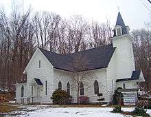A white church with a black roof, pointed windows and a pointed steeple, with woods full of bare trees in the back. There are a few patches of snow on the ground in front.