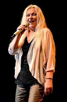 Colored photograph of Toni Willé standing onstage holding a mike. She has long blonde hair and is wearing a black top and off-white full-sleeved top with grey pants.