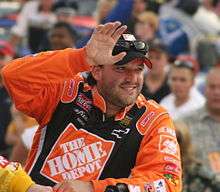 A man in his mid thirties is wearing orange racing overall and his waving to the crowd with his right hand