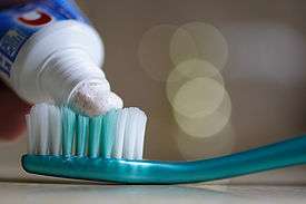 Photo with a blurry background and toothpaste from a tube of toothpaste being applied to the bristles of a toothbrush in the foreground