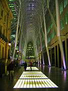 A towering metal gridwork catenary arch forms a gallery over a shiny concrete concourse inset with glowing grids of glass. Beyond the arch, city lights of tall buildings are visible. A four-story historic stone buildign stands to the left; a plain modern building rises out of sight to the right.