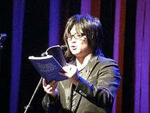  Standing Japanese man reading a book onstage at a microphone