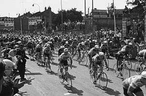 A black and white photograph of a large group of cyclists riding under a starting banner.