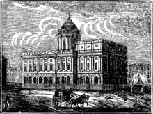 A neoclassical building seen from an angle. It has two storeys and a dome on a high drum. A horse and cart pass in front of it and to the right of the hall is a covered wagon