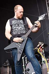 A male in a black sleeveless t-shirt and blue jeans looking at the neck of black flying v guitar while playing it during a performance