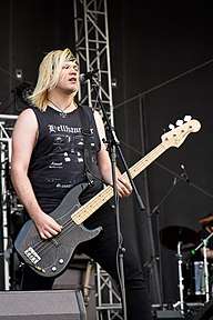 A male with shoulder-length hair, a black sleeveless t-shirt and black jeans playing a bass guitar during a performance