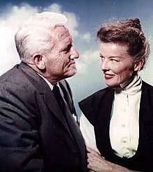 Hepburn is sitting with Spencer Tracy, she age 50 and he age 57, and they are smiling at each other.