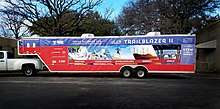 Photo features a 40 ft trailer attached to a pickup truck, with colorful images on the side to show what exhibits the museum houses. These images include: a hurricane for weather, a fighter jet for aerodynamics, an athlete with a J-leg prosthetic for biotechnology, wind turbines on a hillside for energy, and the International Space Station for space.