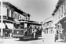 An electric trolley with a man hanging off one side rounds a corner of a street lined by two-story stores and horse-drawn kalesas.