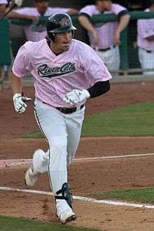 A man in white pants, a pink baseball jersey, and a black batting helmet running down the first baseline.