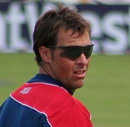 A man wearing sunglasses and navy blue and red English cricket T-shirt looks across over his shoulder at a sports ground. A sponsorship board is visible in the background.