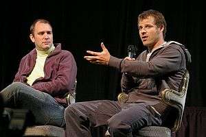 Two men sitting on chairs wearing jeans and hoodies.