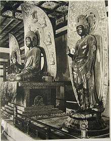 Three-quarter view of a central cross-legged seated statue on a pedestal flanked by two standing statues of almost the same size. The central figure has the palm of his right hand facing forward and that of the left hand upwards with the left hand resting on his knee. The flanking statues show a similar pose with their right hand while their left arm is hanging down. All three statues have halos in their back which are decorated with small cross-legged seated statues.