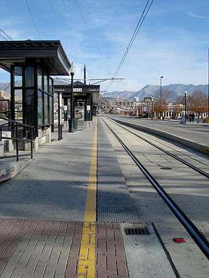 An empty island platform station located next to the tracks in the median of the street with canopies visible. Businesses line the street with mountains visible in the distance.