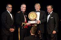 A ceremony in which Trump receiving the 2015 Marine Corps–Law Enforcement Foundation's annual Commandant's Leadership Award. Four men are standing, all wearing black suits; Trump is second from the right. The two center men (Trump and another man) are holding the award.