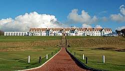 A golf course. In the background is the Turnberry Hotel, a two-story hotel with white façade and a red roof.