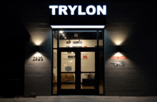 A dark storefront facade with the words "TRYLON" in illuminated white capital letters above the double glass doors. On either side of the doors, lamps cast triangles of light upward and downward to form the shape of an hourglass or bowtie rotated one quarter turn.