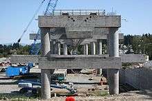 An unfinished concrete structure on a series of columns. A mobile crane and other construction equipment can be seen below the structure.