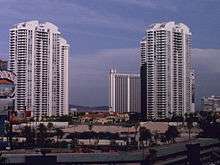 Distant ground-level view of four identical 40-story buildings; each structure has a white concrete facade with balconies at every floor level and a rounded roof.