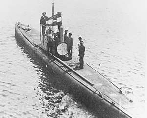 A submarine floats on the surface with its crew standing on the deck and conning tower. The naval ensign of Austria-Hungary can be seen flying from the submarine's conning tower and the main entry hatch of the boat is open.
