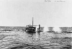A photo taken from near the waterline of a submarine diving. Nearly the entire boat is underwater, with just the conning tower remaining above the surface. Spouts of water can be seen erupting on the surface from the submarine's air vents as it dives.