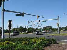 Several cars pass through a signalized intersection viewed from behind planted shrubs. Small green signs on the traffic light pole masts indicate the roads are named Ocean Highway and Worcester Highway.