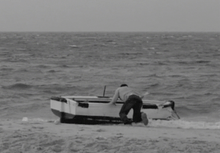 A black and white image showing a man as he pushes a rowboat over the sand and tries to get it into the choppy water. The boat, which is white on top and black on the bottom, is just entering the surf from the side.