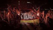 A close-up shot of fans inside a stadium during a concert, many of whom have their arms in the air. A transparent layer on top shows a concert stage with a curved video screen showing black-and-white images of musicians performing, with solid red and yellow colors below.
