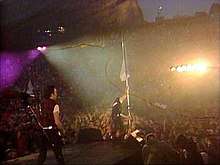 A shot of a concert stage from the back at dusk. On the left, the Edge plays guitar. In the center, Bono, at the edge of the stage, holds up a white flag near audience members, several of whom help him support the flag. In the center of the audience are bright lights aimed in the direction of the performers. Behind the audience are large red cliffs. Red streaks of light are visible throughout the image