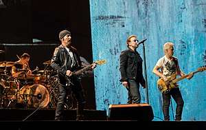 Four men on a stage, all wearing black clothing. Two are playing guitars, one is sitting behind a drum set, and one is singing into a microphone stand.