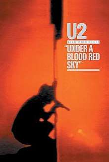 A red background encompasses most of the shot. In the lower left corner is a shadowy silhouette from the side of a man crouching down, holding a microphone to his mouth and a flag into the air. Written in grey text in the upper right corner are the words "U2 – Live at Red Rocks: Under a Blood Red Sky".