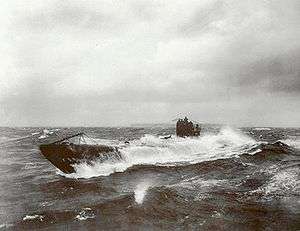 A black and white image of a submarine breaking through a wave in rough water.