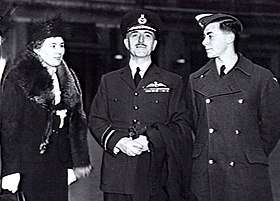 Three-quarter outdoor portrait of moustachioed man in military uniform with peaked cap and pilot's wings on left breast pocket, flanked by woman in hat and fur coat, and young man in military great coat and forage cap