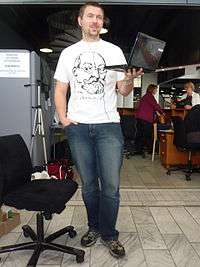 In a cavernous convention centre of industrial design demarcated with temporary office partitions, a man with a goatee and close-cropped brown hair and earphone buds is standing, holding an open laptop in his left hand. His right hand, except for his thumb, is in the pocket of his blue jeans, and he wears a white T-shirt with the signature and three-quarter profile outline of Esperanto founder L.L. Zamenhof, a friendly, full-bearded but balding man with a prominent nose and round glasses. Behind him is a circular staircase; to his left two women are in conversation.