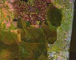 A color satellite image of the northern Everglades showing green chunks of Everglades surrounded by white settlement areas of the South Florida Metropolitan Area to the east and red agricultural fields in the Everglades Agricultural Area to the north