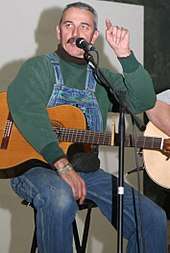 A grey-haired man with a mustache, wearing a green shirt and bib overalls, talking into a microphone while holding a guitar