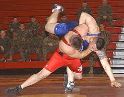 Two men wrestling in a gymnasium, watched by a group of uninformed soldiers