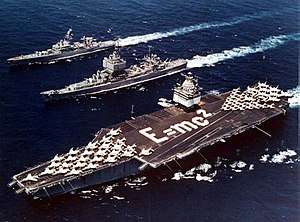 Three ships sale next to each other, including an aircraft carrier which has jets arranged as an arrow and the phrase E=MC2 written with sailors on deck