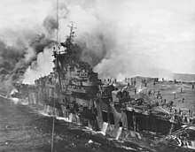 Black and white photograph of a World War II-era aircraft carrier on fire. The portion of the ship rear of its island has been consumed by a huge fire. People with large hoses are visible on the ship's flight deck.