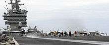 Three gray F/A-18 Hornet strike fighter aircraft line up across the frame for catapult launches from an aircraft carrier's deck. Support staff is seen on the deck throughout, while exhaust can be seen from the engines of the aircraft on the right.