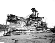 A stern view of an incomplete ship sitting in a drydock with the top of the main turret open, awaiting armor plates.