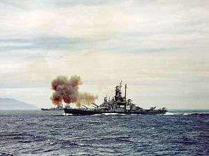 Color photo of a warship at sea. Smoke is rising from the bow of the ship, and land is visible in the background.