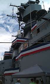 A photograph of the ship's superstructure as seen from deck level. The bridge, radar mast, and a phalanx gun are visible.