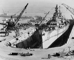 A shipyard with a large dry dock occupied by a massive gunship. Crewmen can be seen on the battleship's deck, while dock equipment such as cranes and trucks can be seen lining the sides of the drydock. In the distance a pier can be seen, while two smaller ships are visible in the background of the image.