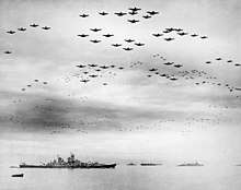 Black and white photo depicting a large number of aircraft flying in formation above a group of World War II-era warships