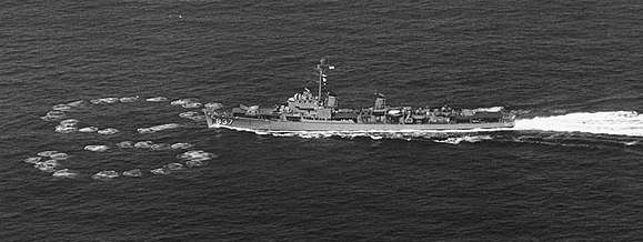 A destroyer moving to the left; on the water just ahead of her are two rings of small circular waves, one to either side of her path. The rings nearly meet directly ahead of her and are each about half her length in diameter.