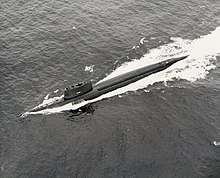 A surfaced submarine is seen from above and to port making high speed, with a long wake around and behind.