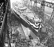 A large warship, still missing most of its superstructure, sits in a dry dock, awaiting its launch. The ship is draped in a large banner and surrounded by crowds of spectators; a huge gantry towers over the ship.