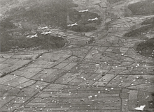 Aerial photograph taken from a high angle. Six twin-boomed aircraft are flying in two formations of three, while below them a large number of parachutes are suspended in the air over a cultivated field